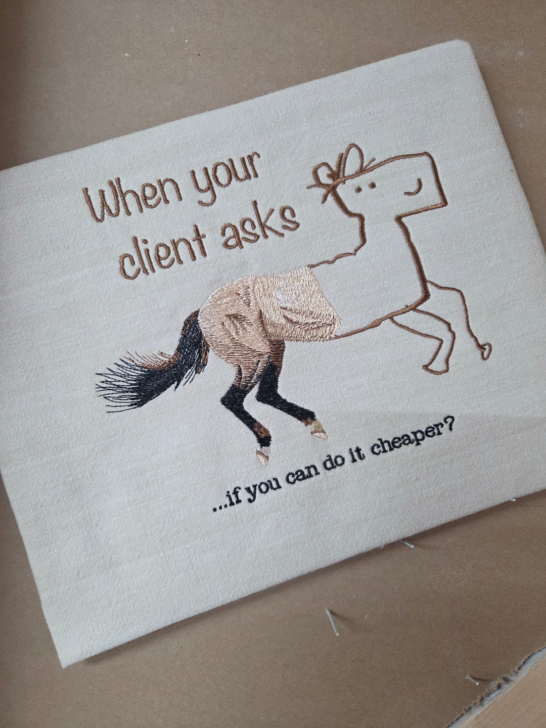 A square white fabric napkin or coaster featuring a whimsical embroidered horse and the text 'When your client asks if you can do it cheaper