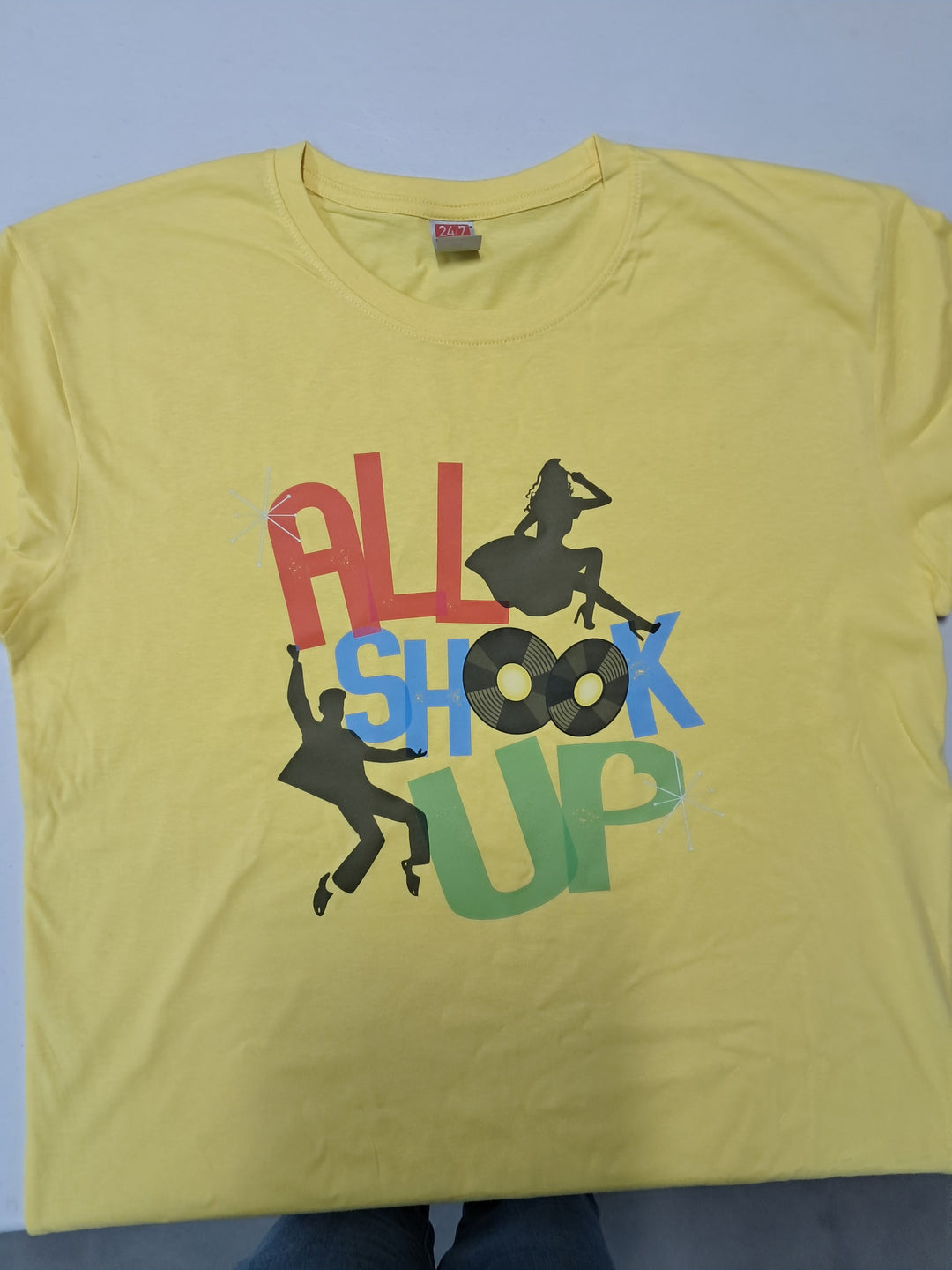 A yellow short-sleeve t-shirt with a colorful retro-style graphic reading 'ALL SHOOK UP' in red, blue, and green, accompanied by black silhouettes of people dancing, laid flat against a light gray background.
