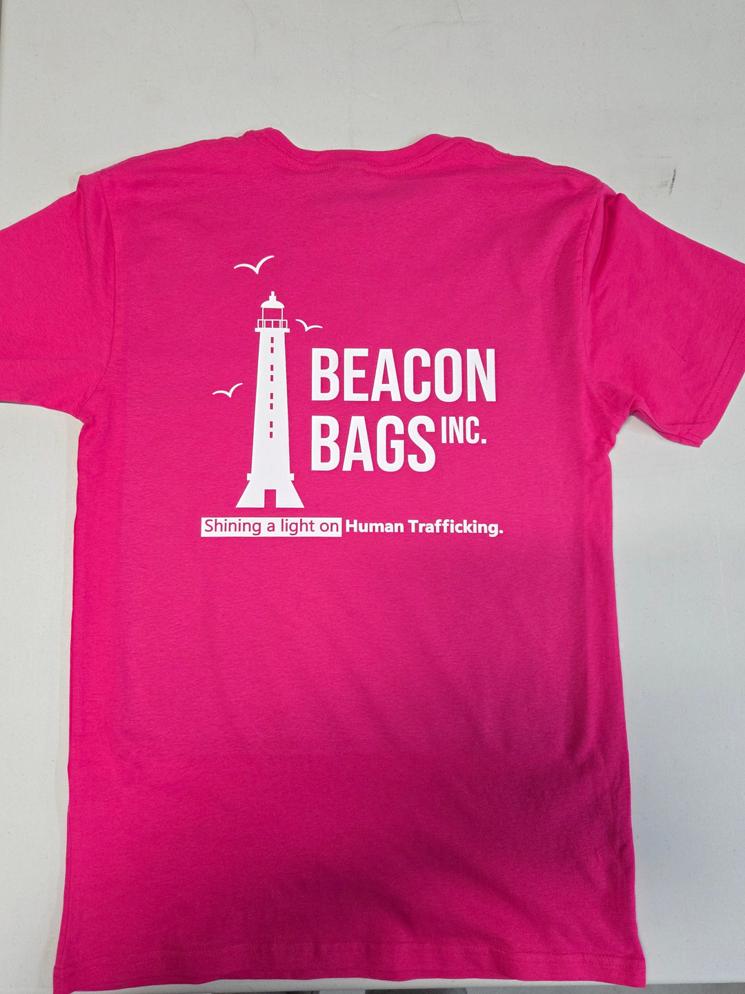 A bright pink short-sleeve t-shirt laid flat on a light gray surface, showing the back side with a white graphic of a lighthouse, birds, and the text 'BEACON BAGS INC.' and 'Shining a light on Human Trafficking.