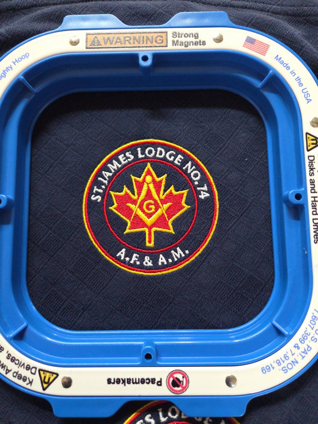 A close-up of a blue and white square magnetic garment steamer holding a dark navy fleece fabric with a circular embroidered St. James Lodge No.