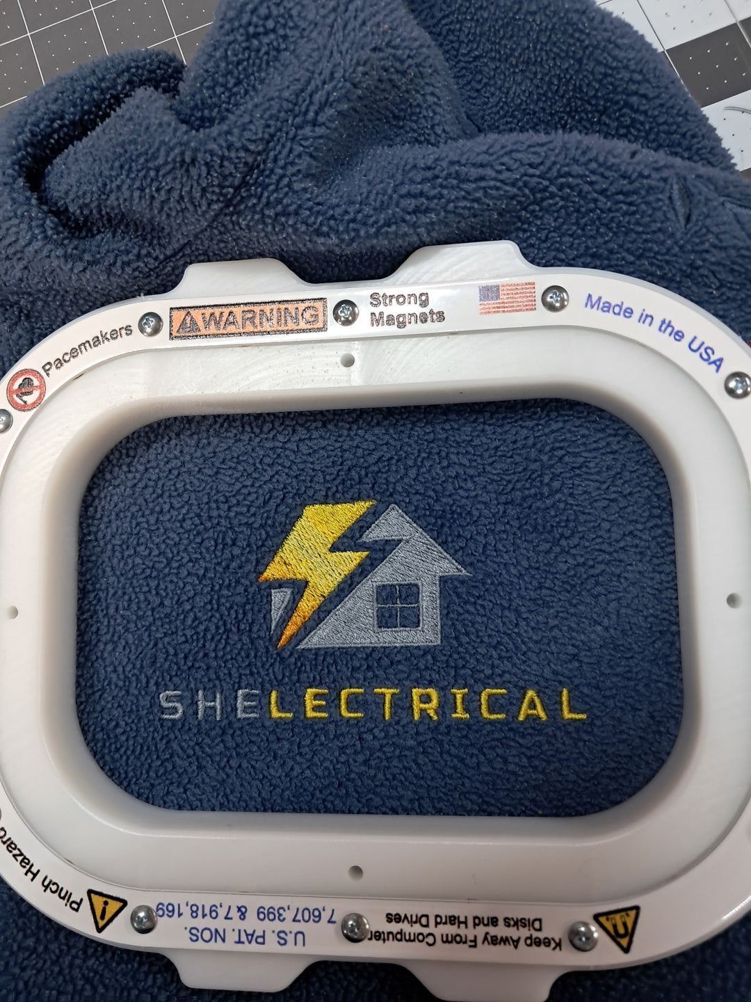 A close-up of a navy blue fleece-lined patch or badge with a white plastic outer frame. The center features a textured navy blue background with a yellow lightning bolt and gray house logo above the word 'SHELECTRICAL' in yellow.