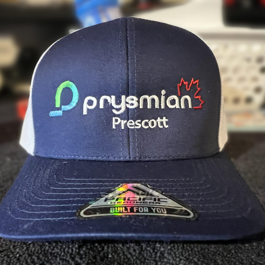 A navy blue and white trucker hat with 'prysmian Prescott' embroidered on the front, featuring a colorful Pacific Coast brand sticker on the brim.